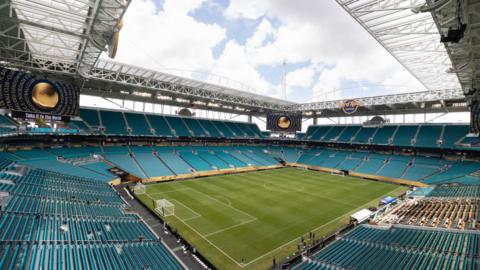 Empty football stadium, with green pitch and rows of turquoise seats and roofing.