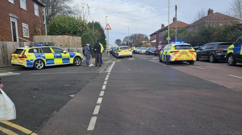 A number of police cars are parked around a junction. Two men speak to a police officer