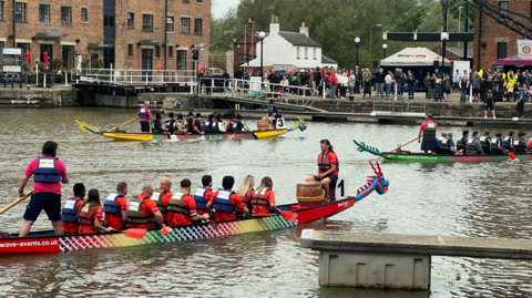 Three boats with 10 paddlers each are on the water in a city's docks. They are all wearing lifejackets and there are buildings visible, ringing the water's edge, with dozens of spectators watching too