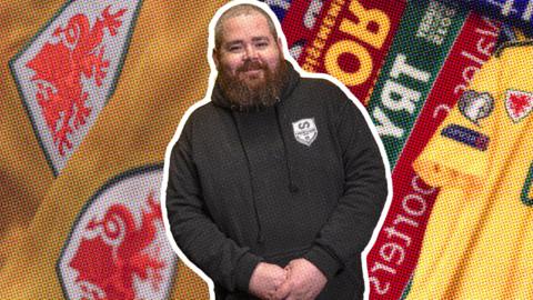 Mid-thirties man with beard and black jumper in front of football shirts and scarves