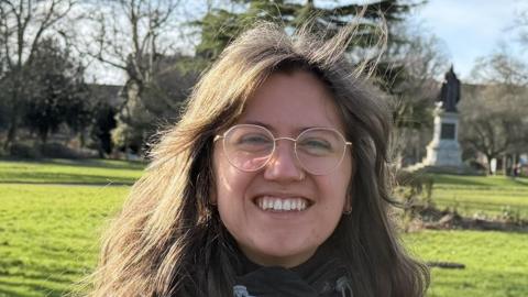 Miriam Stobart has brown hair and round gold glasses. She is smiling at the camera and has a nose ring. She is sitting on a bench in front of a park with a statue in the background. It is a sunny day. 