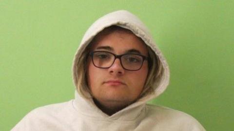 A police picture of Jessica Garnham-Burton. She is wearing a white hooded jumper with the hood up. She has blonde hair and black glasses. 