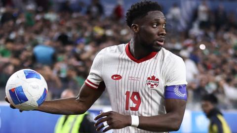 Alphonso Davies of Canada, in the team's largely white kit with red detail, holds a football in his right hand before taking a throw-in