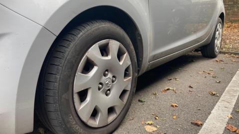 The close-up of the side of a silver vehicle focusing on the tyre. The centre of the tyre is silver and the rubber is black. The car is parked in a parking bay, with a white line on its right.