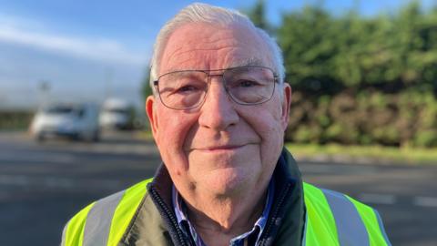 A man with short grey hair and glasses, wearing a high-vis jacket, smiles at the camera. He is standing in front of a crossroads as vehicles wait to move off.