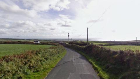 The road, it is a country lane, there are fields around and houses dotted across the landscape.