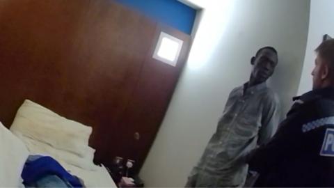 Deng Chol Majek is standing next to a police officer in his hotel room.