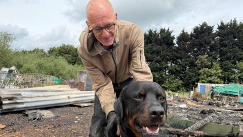 Martin Taylor with his hands around his Rottweiler dog who is standing in front of him. Martin has a shaved head and is wearing glasses and a beige coat with dark trousers