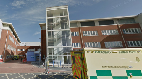 Nondescript brown hospital building with large windows with green ambulance in front.