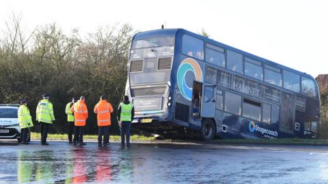 The Stagecoach bus is across a grass verge with its front wheels in a ditch and trees and houses in the background. Police are at the scene in hi-vis jackets and there are more people in orange jackets. The ground is wet.