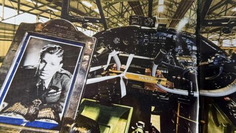 Framed picture of Guy Gibson with a pair of his cloth wings inset. The picture has been placed inside the cockpit of a Lancaster bomber.