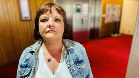 Woman - Lynn Winstanley - looking at camera inside a council building, she is wearing a white v-neck top under a light blue denim jacket, and a necklace.