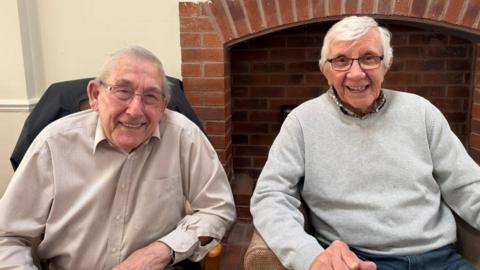 The photo shows two elderly men smiling at the camera. They both have glasses and grey hair. The one on the left is wearing a brown checked shirt. The one on the right has a grey jumper with a shirt underneath. They are sitting in armchairs, in front of a fireplace. 