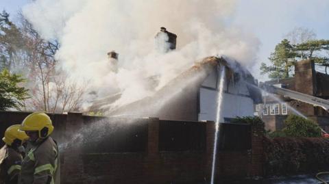 Thatched cottage with its roof on fire and tick smoke billowing out. Two firefighters are seen to the left and several hoses are spraying water onto the roof.
