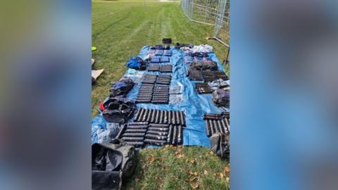 Dozens of packets of drugs laid out on a blue groundsheet in a field.