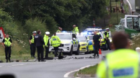 The scene of a crash on the A1 near Causey Park. A mass of police officers are standing in the road, which has been cordoned off, with police cars parked with their blue flashing lights on. Vehicular debris is strewn across the road.