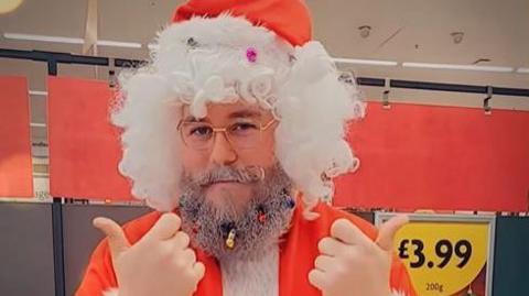 A man dressed as Father Christmas using sign language in a supermarket with a price sign behind him.