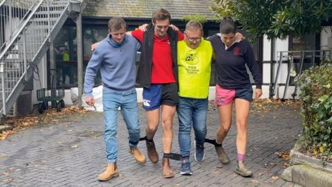 Three men and one woman (right), with their legs tied together with black elastic so they are three-legged. They have their arms around each other.