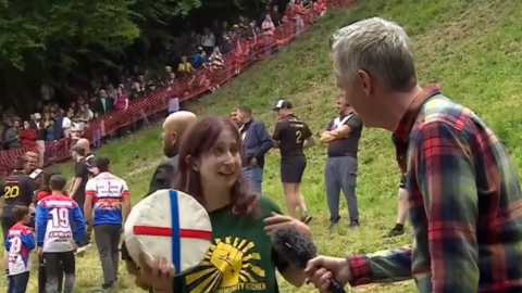 Woman in green top holds up a wheel of cheese while being interviewed, people in comfortable clothing walking behind her
