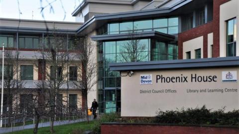 The photograph shows the outside of Phoenix House, Tiverton. In the foreground is a sign that reads "Phoenix House, District Council offices". The building behind the sign has a glass entrance and the building appears to be three to four storeys high.