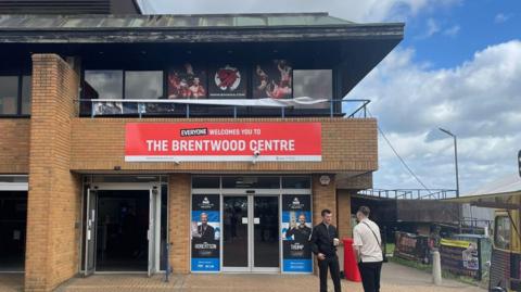 Two people standing outside the main entrance doors at the Brentwood Centre. There is a red banner above the door welcoming people.