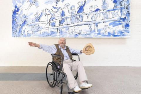 Quentin Blake, sat in a wheelchair, wearing a light blue shirt and brown waist jacket and holding a tan hat with his arms outstretched under a large blue and white mural. 