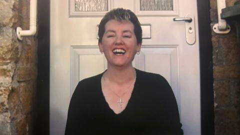 A woman with short hair in front of a white front door. She has small hoop earrings and a cross pendant on, and is grinning broadly. She is wearing a black top.