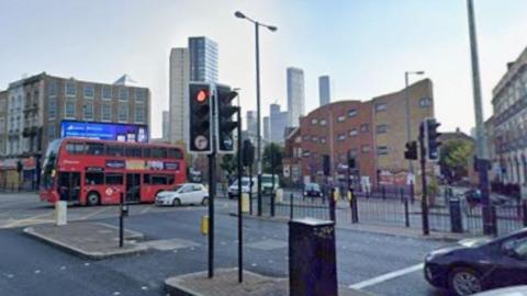A busy road junction in east London, there are cars and red buses on the roads and high-rise commercial and residential buildings in the background.