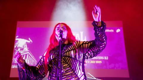 A woman with long red hair on stage in a spider's web outfit. There is a large screen behind her. 
