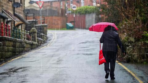 A woman walks through Mayfield in Staffordshire in the rain. She is seen from behind and is carrying a red umbrella