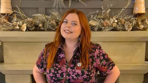 Rachel Clark standing in front of a stone fire place in a pink and blue floral top 