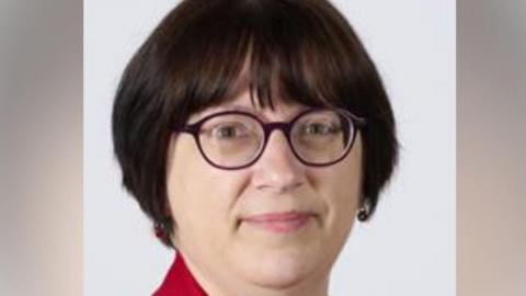 A close up image of a professional portrait image of Ruth. She has a short black bob and full fringe. She has round purple glasses and wears a red shirt. She is smiling slightly at the camera.