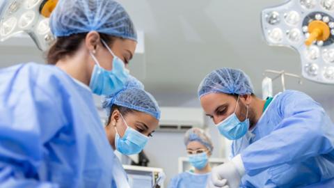 A surgical team in an operating room performs a procedure on a patient. The team is wearing surgical gowns, gloves, and head coverings, working around an operating table with medical instruments.