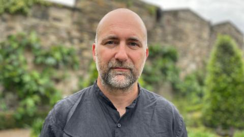 Simon Francis from the End Fuel Poverty Coalition is picture with a blank expression standing in a garden wearing a great shirt 