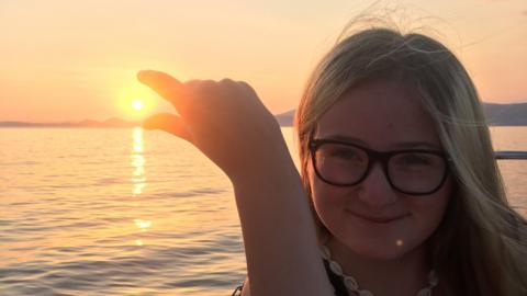 Lucy Curtis holds her fingers up to look as if she is pinching the sun as it sets while standing next to a lake. She has blonde hair and glasses.