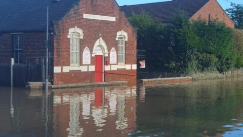 A brick building with a red door and arched windows outlined in white face on to a road which is underwater.