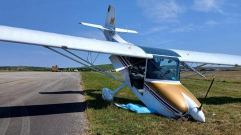 The photograph shows a small, high-wing light aircraft just off the edge of a runway at Land’s End Airport. The aircraft's nose is buried in the ground, causing the tail section to point sharply upwards into the air. The front of the fuselage appears to have dug into the grass, leaving the aircraft in a nose-down position. 