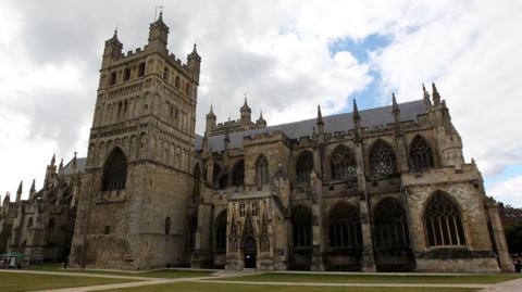 An outside shot of Exeter Cathedral. It shows all the architecture and the cathedral green. There is the main building and the tower which is at the heart of it. The weather is cloudy with a blue sky behind them. 