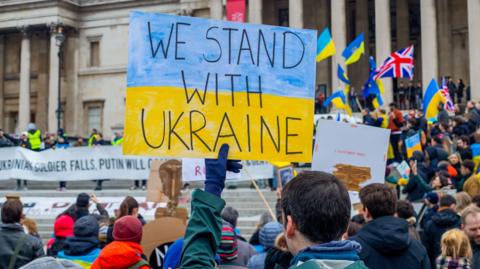 A protester holds a sign in the colours of the Ukrainian flag reading: We stand with Ukraine. He is in a crowd of people who are holding Ukrainian flags and Union Jacks. 