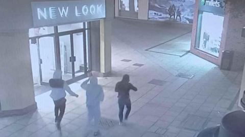 A still from CCTV showing three people in hoodies walking away from the camera with shops either side of them. One is in a white and black tracksuit, another is in all grey and another is in all black.