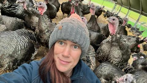 Kate Postance of Postance Poultry takes a selfie in the the purpose-built tunnel housing her turkeys.  The turkeys can be seen behind her.  Kate has long brown hair and is wearing a grey, woollen hat and a blue fleece.
