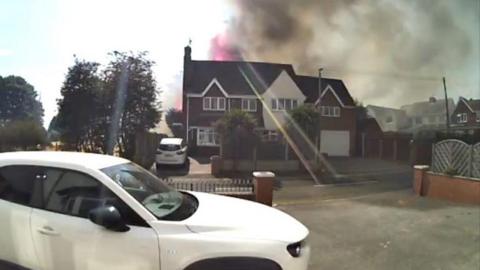 Smoke and flames are rising above a house with a white car parked on the drive.