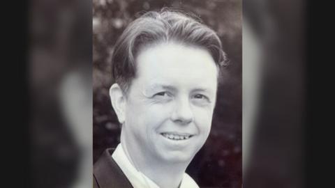 A black and white photograph of Simon Scott smiling at the camera while wearing a white shirt and dark jacket.