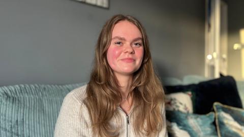 Mollie Penfold who has brown long hair and is wearing a grey quarter zip wooly jumper sitting on the sofa of her living room
