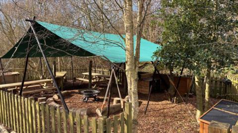 The woodland classroom at Hauxley Nature Reserve. It is an area surrounded by a wooden fence. It has a canvas cover over a central area with a metal firepit, picnic benches and other seating.
