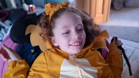 Addy smiles and looks away from the camera. She has curly hair and wears a yellow and white onesie or fleece, with a yellow bow in her hair. 