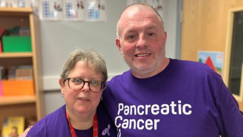 A man and a women stood next to each other smiling. They are both wearing a purple Pancreatic Cancer UK T-shirt. They both have grey hair and the women is wearing glasses.