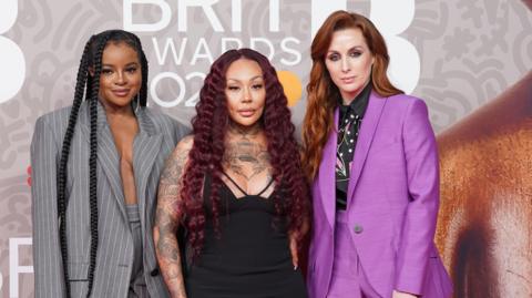 Left to right: Keisha Buchanan wearing a grey and white striped suit. Mutya Buena wearing a black dress. She has tattoos on her arms and chest. Siobhan Donaghy wearing a purple suit with a black blouse underneath. They are standing together looking at the camera. 