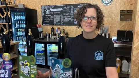 A woman with short brown curly hair and glasses wears a black t-shirt while standing behind a bar with three beer taps in front of her. Behind her are several fridges with bottled drinks inside them.