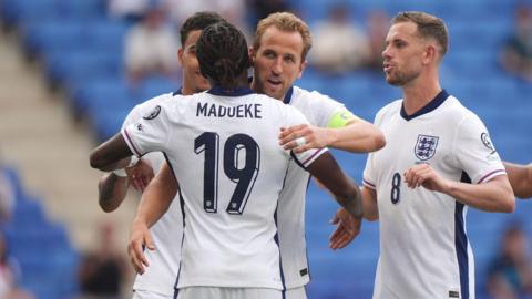Harry Kane is about to hug his teammate, Noni Madueke. They are wearing white England football shirts with navy blue accents on them. Harry Kane is wearing a bright green captain's armband. Next to Harry is Jordan Henderson, moving in for a hug.